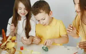 children-paint-eggs-mother-teaches-children-sitting-white-table Moment d’activités éducatives : des enfants et une femme peignent des œufs de Pâques, stimulant leur créativité et leur motricité fine.